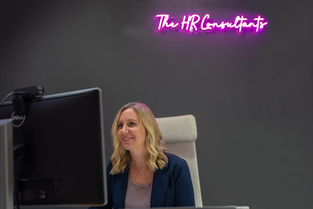 Person working at desk computer with Neon Sign behind them, reading 'The HR Consultants'